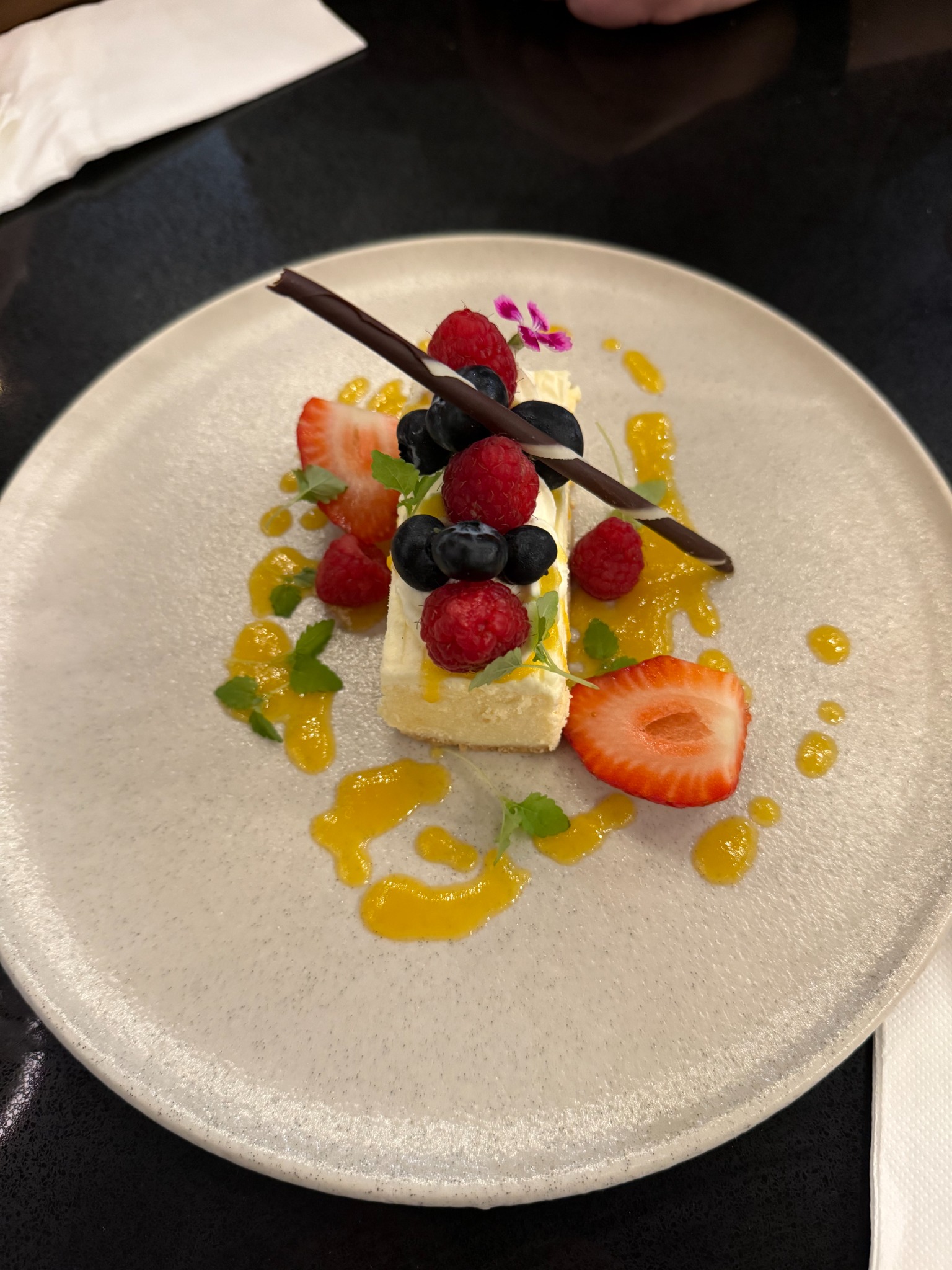 A rectangular, pale dessert with berries, chocolate decoration and yellow sauce.